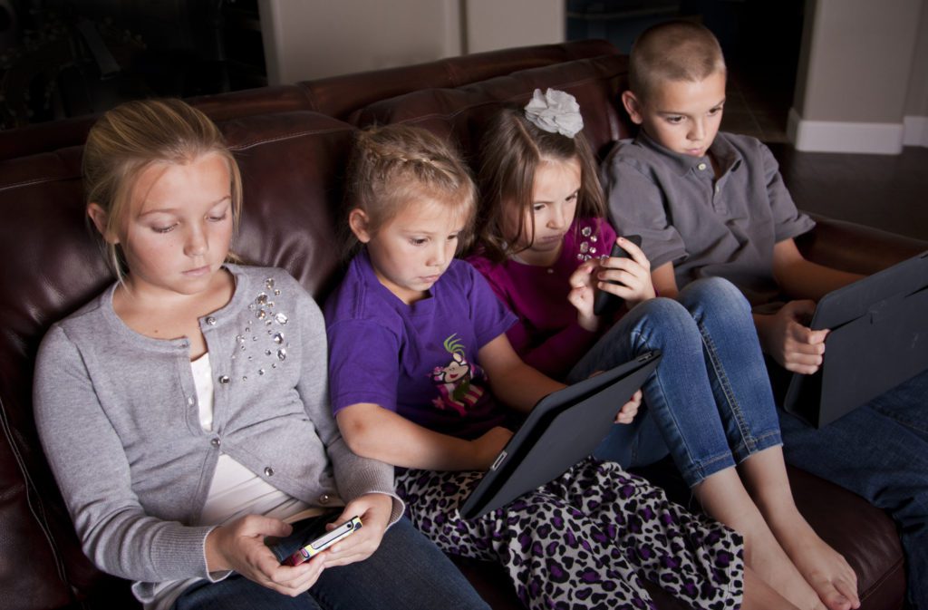 A row of four young children sitting on a leather couch, fixated on the mobile devices they are each holding.